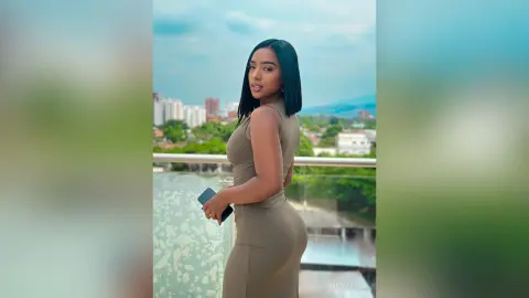 Young woman in a tight olive green dress posing on a balcony overlooking a city skyline under a blue sky.