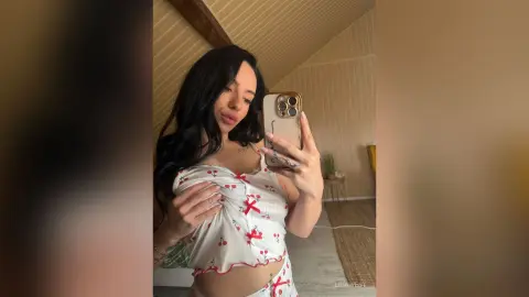 Young woman with long dark hair wearing a white floral print crop top taking a mirror selfie in a cozy room.