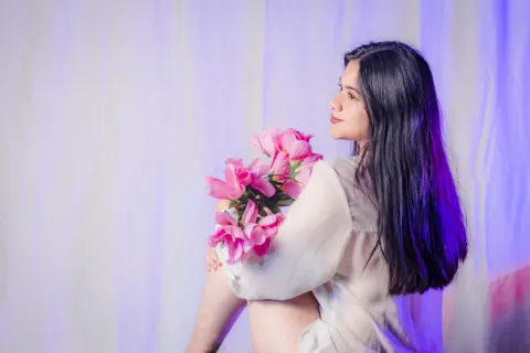 Young woman in a white blouse holding pink flowers against a soft purple backdrop in a dreamy mood.