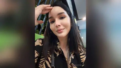 Smiling young woman with long dark hair wearing a tropical print blouse in a bright cafe setting.