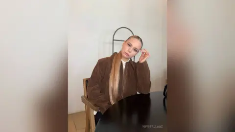 KrystalLaaz sits at a black table wearing a brown jacket and looking at the camera with her long blonde hair down.