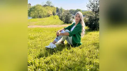 Blonde woman in a green blazer and white sneakers relaxing on a sunny green grass field.