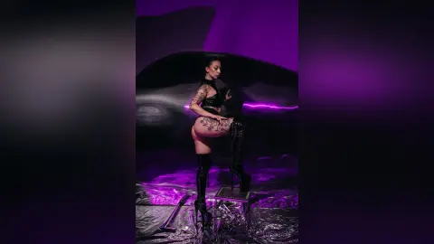 Glamorous woman in black bodysuit and lace-up boots posing in a moody purple studio setting.
