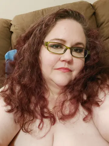 Smiling woman with curly red hair and green glasses relaxing indoors in a cozy setting.