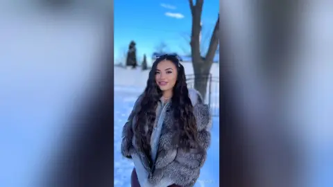 Smiling woman in a cozy faux fur coat enjoying a sunny winter day in a snowy landscape.