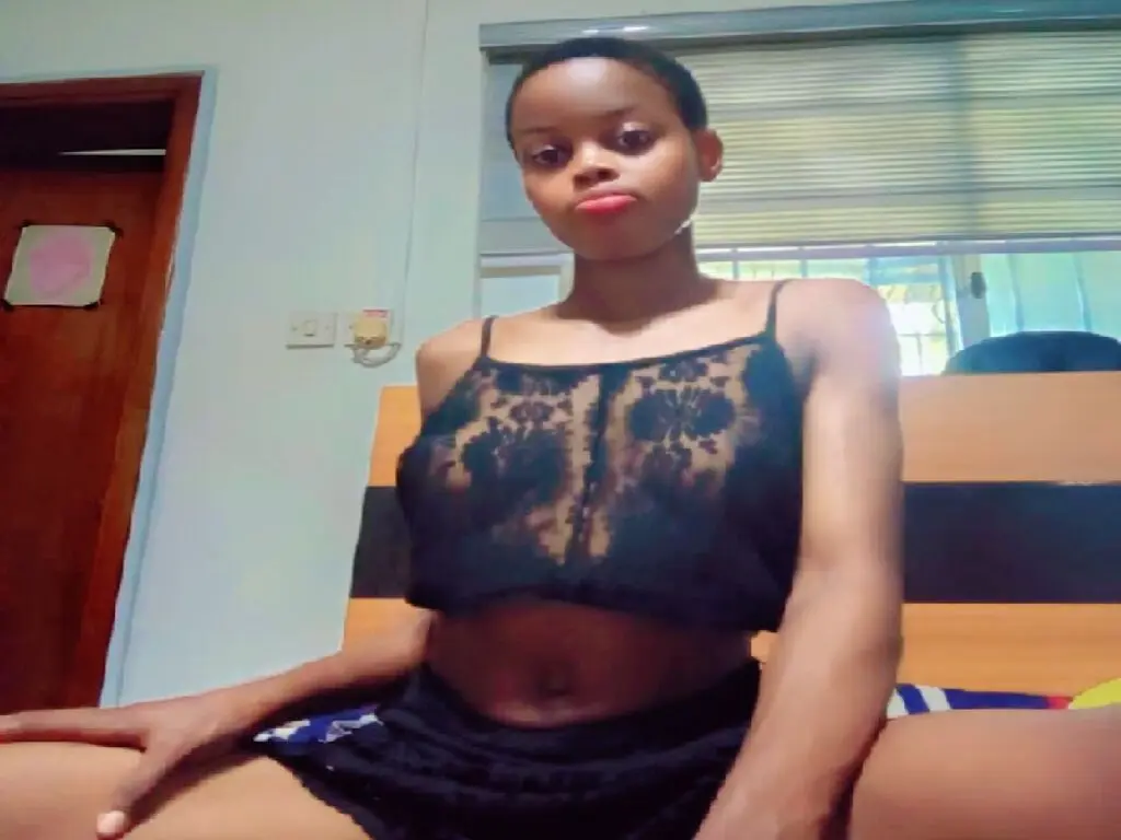 Young woman in a black lace crop top sitting relaxed in a sunlit bedroom.