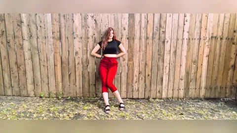 Stylish woman in a black t-shirt and red trousers posing against a rustic wooden fence in natural light.