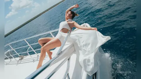 Elegant woman in a flowing white dress relaxing on a boat in the turquoise ocean under a sunny sky.