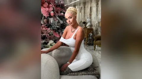 Glamorous blonde woman in a white cocktail dress sitting in an elegant, floral-decorated luxury living room.