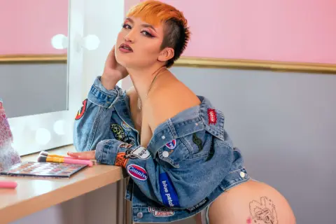 Young woman with orange hair in a denim jacket posing confidently in a bright makeup studio.
