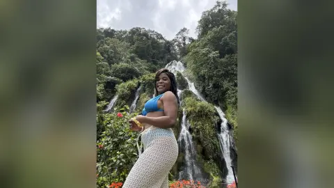 Smiling woman in a blue bikini and white crochet skirt relaxing by a lush tropical waterfall.