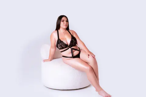 Curvy woman in black lace lingerie sitting on a white faux fur chair against a clean studio background.