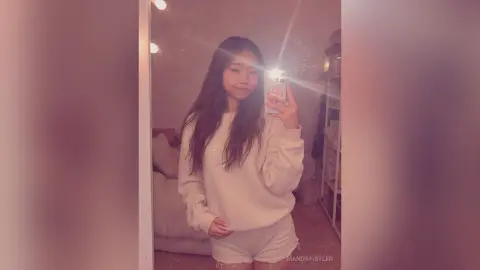 Young woman in a cozy white sweatshirt taking a casual mirror selfie in a dimly lit bedroom.
