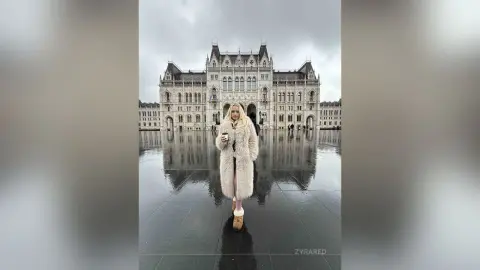 Blonde woman in a cozy faux fur coat standing before the Hungarian Parliament Building on a rainy day.