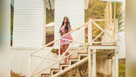 Miah stands on a white wooden deck wearing a red and white checkered sundress with thin straps.