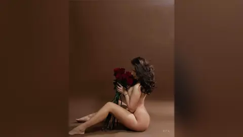 Nude woman sitting against a brown background holding a bouquet of red roses in a romantic, moody setting.