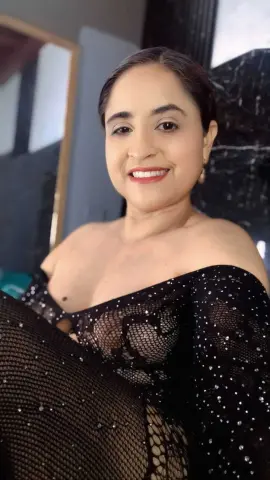 Smiling woman in a black sequined mesh off-the-shoulder dress posing in a modern indoor setting.