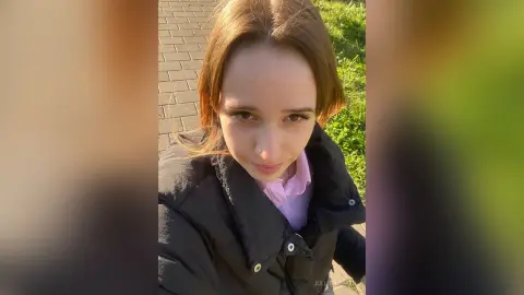 Young woman in a black puffer jacket and pink shirt smiling outdoors in sunny garden setting.