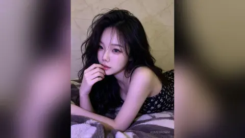 Young woman with long dark hair in a black floral top relaxing on a cozy bed in a soft, moody indoor setting.