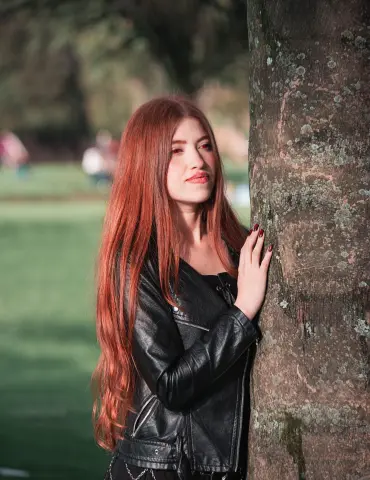 Redheaded woman in a black leather jacket leaning against a tree in a sunny park.