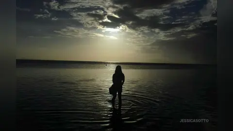 Silhouette of a woman standing in calm ocean water under a dramatic sunset sky at dusk.