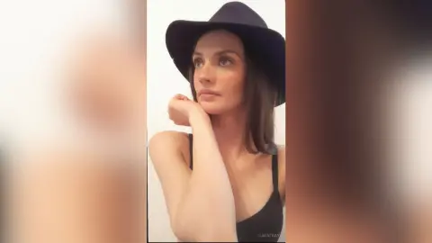 SaraTray wears a black hat and a black bra, posing with her hand under her chin against a plain white background.