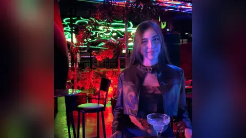 KateGale smiles while wearing a black leather jacket and a dark top, holding a margarita in a dimly lit bar setting with neon lights and plants.