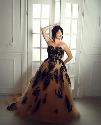 Elegant woman in a black lace strapless ball gown posing by a bright window in a classic white room.