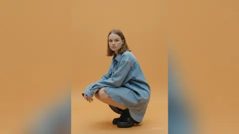 Young woman in an oversized denim shirt and black boots posing against a warm tan background.