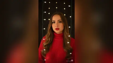 Young woman in a cozy red turtleneck sweater posing under warm fairy lights in a festive, moody setting.