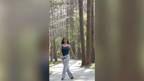 Smiling woman in a blue tank top and grey sweatpants walking through a sunny forest path.