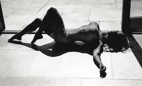 Moody black and white silhouette of a woman in a textured bodysuit lying on a sunlit stone patio.