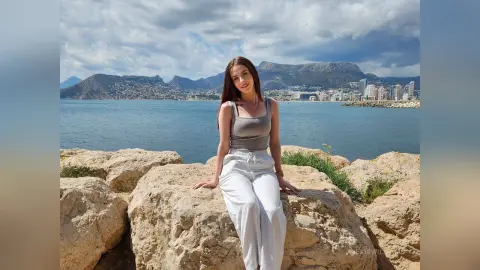 Young woman in a grey tank top and white trousers sitting on rocks by the sea under a cloudy sky.