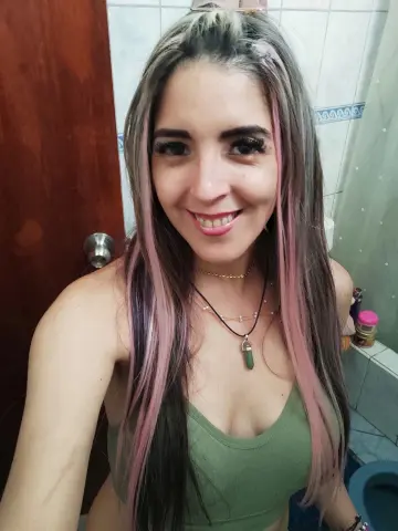 Smiling woman with long blonde and pink hair wearing a green tank top in a bright bathroom setting.