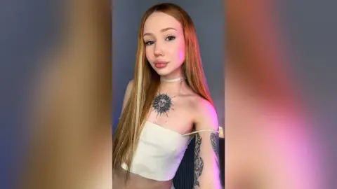 Redheaded woman in a white tube top with sun chest tattoo posing against a studio background.