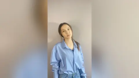 Young woman in a light blue button-down shirt posing against a neutral wall in a minimalist indoor setting.