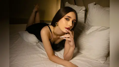 Glamorous woman in a black slip dress and gold jewelry relaxing on a white bed in moody lighting.