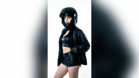 Edgy woman in black leather jacket and biker helmet posing against a white background with a cool, moody vibe.