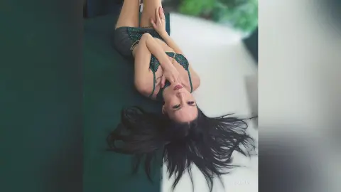Young woman in a green lace bodysuit lying on a white surface with long dark hair in a moody pose.