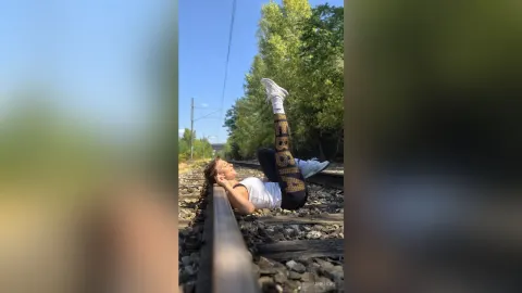 JaneHope — JaneHope lies on railroad tracks with her legs raised toward the sky, wearing a white tank top and black patterned leggings.