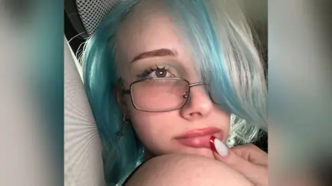 Young woman with blue hair and silver glasses posing with a dreamy expression in a cozy indoor setting.