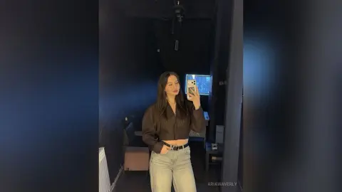 Young woman in a brown long-sleeve crop top and light denim jeans taking a mirror selfie in a moody dark room.
