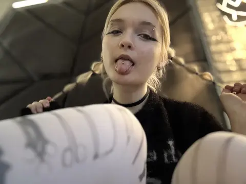 Young woman with blonde hair and dark eyeliner sticking out her tongue in a moody indoor setting.