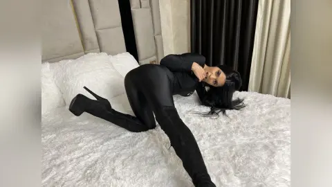 Woman in black bodysuit and high heel boots lounging on a white fluffy bed in a cozy bedroom setting.