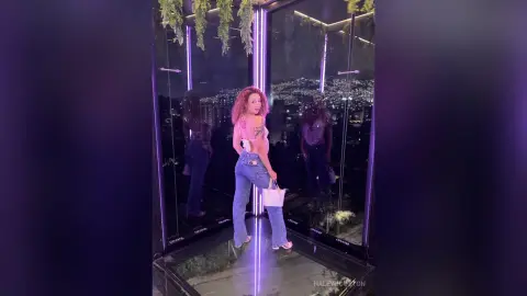 Young woman in a white crop top and blue jeans posing in a modern rooftop lounge with purple neon lighting.