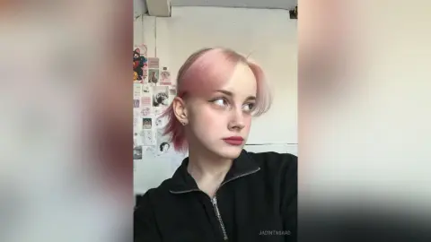 Young woman with pink hair and black zip-up sweater looking away in a bedroom setting with a moody aesthetic.