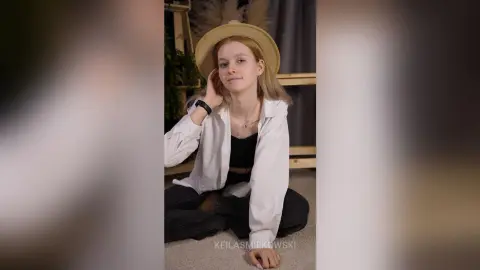 Smiling young woman in a white button-down shirt and wide-brimmed hat sitting on the floor in a cozy room.