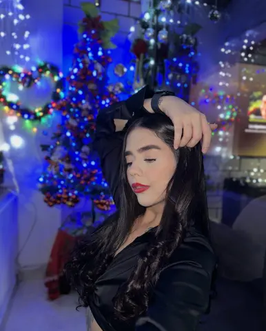 Nomohah_sainz poses with her hands in her long dark hair, wearing a black shirt against a background of blue and colorful holiday lights.