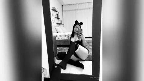 Young woman in black lingerie and stockings taking a selfie in a bedroom mirror with a moody aesthetic.