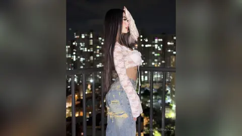 Young woman in a white lace crop top and denim jeans posing on a balcony overlooking city lights at night.